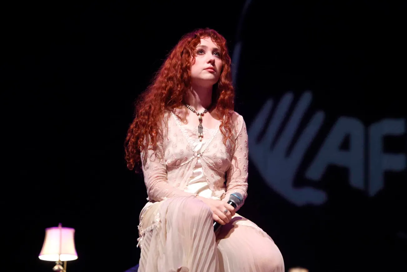 Chappell Roan sits on a dark stage wearing a cream outfit. She has long red curly hair.