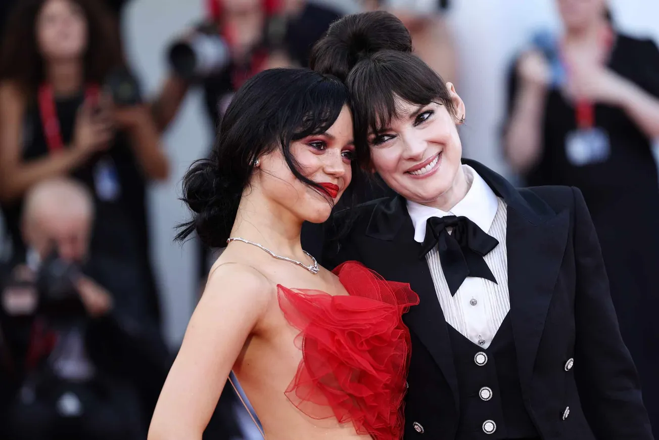 Jenna Ortega and Winona Ryder on the red carpet togehter. Their heads are leaning in towards each other and they are both smi