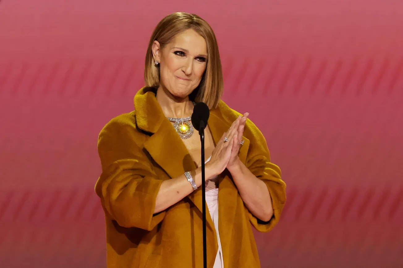 Celine Dion wears a burnt orange coat and stands on stage smiling with her hands together at the 2024 GRAMMYs