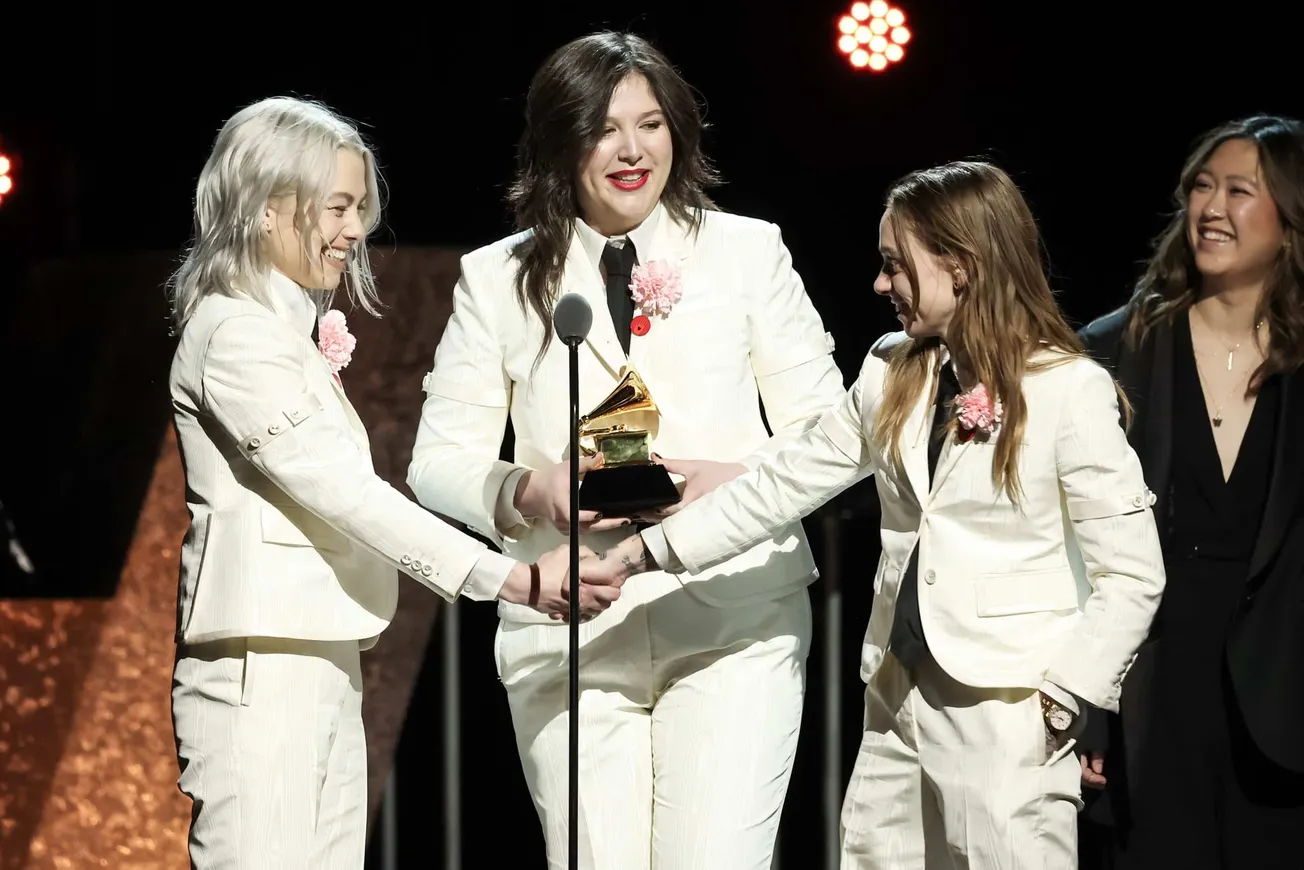 Phoebe Bridgers, Lucy Dacus and Julien Baker all in white suits holding hands and looking at each other smiling as they win a