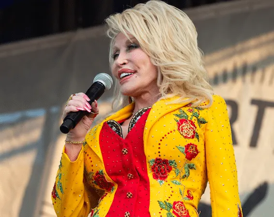 Singer Dolly Parton standing on stage with a microphone, smiling. She wears a yellow jacket with red flowers on them.