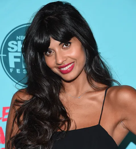 Jameela Jamil smiles at a red carpet event. She has long black hair and wears a black dress with thin straps.