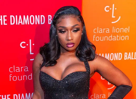 Megan Thee Stallion posing with one arm on her hip at a red carpet event. She wears a fitted, halter-neck black dress and elb