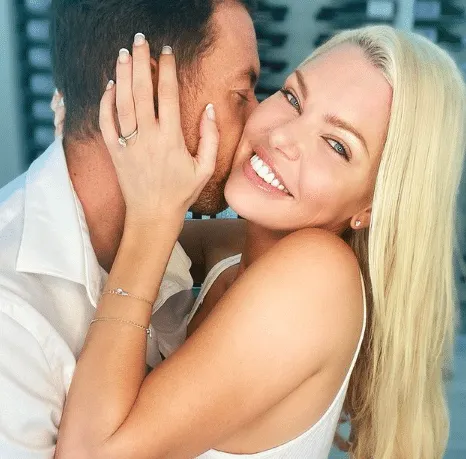 Sophie Monk smiles while Joshua Gross kisses her cheek. She has long blonde hair and wears a diamond ring on her finger as he