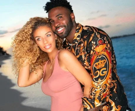 Jena Frumes wears a fitted peach dress that shows her baby bump. Jason Derulo caresses her belly and smiles at the camera. Th