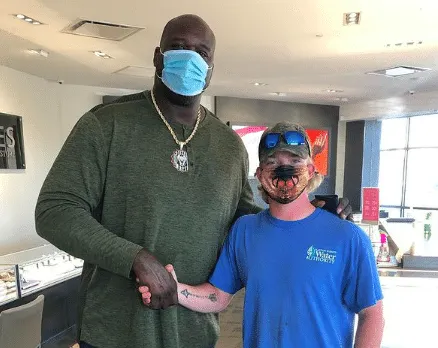 Shaquille O'Neal wears a mask and shakes hands with a man who is also masked and wearing a blue t-shirt.