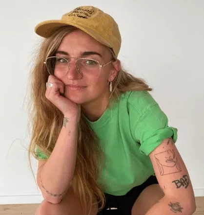 Photo of G Flip wearing a green tee, brown cap and glasses. One had is on their kneww and the other sits under their cheek. T