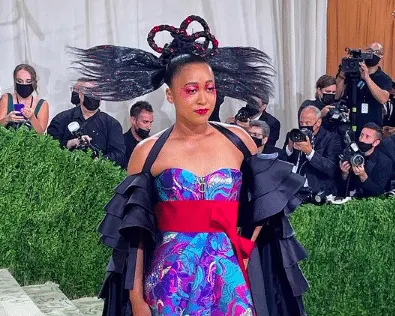 Naomi Osaka at the 2021 Met Gala wearing a black and purple Louis Vuitton dress with ruffles, a red belt.
