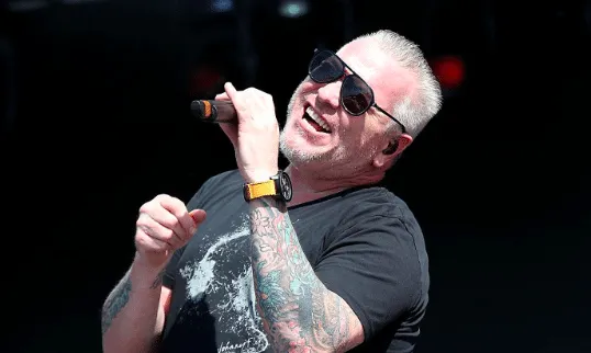 Steve Harwell of Smash Mouth performing on stage in 2017. He's dressed in black and wearing sunglasses. He's holding a microp