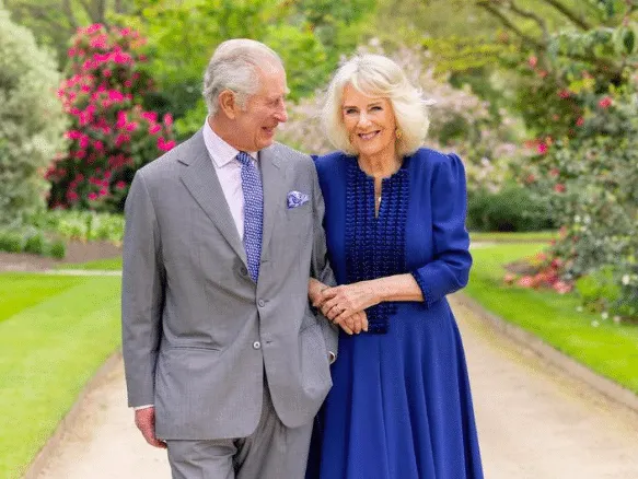 Portrait of King Charles and Queen Camilla smiling as they walk with linked arms through an outdoor, green space.
