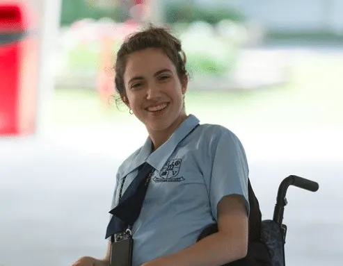 Hannah Diviney as her character Norah Lipsick in 'Audrey'. She is wearing a school uniform and sitting in a wheelchair, laugh