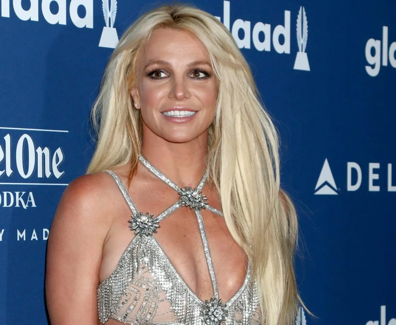 Britney Spears smiles at a red carpet event. She has long blonde hair and wears a silver dress with a bikini style top. Today