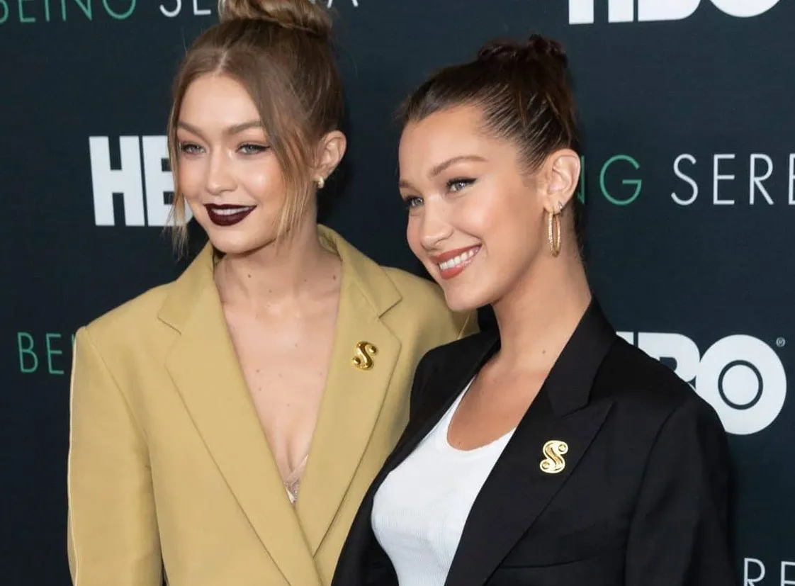 Gigi Hadid and Bella Hadid smile together at a red carpet event. Gigi wears a camel suit and Bella wears a black blazer with