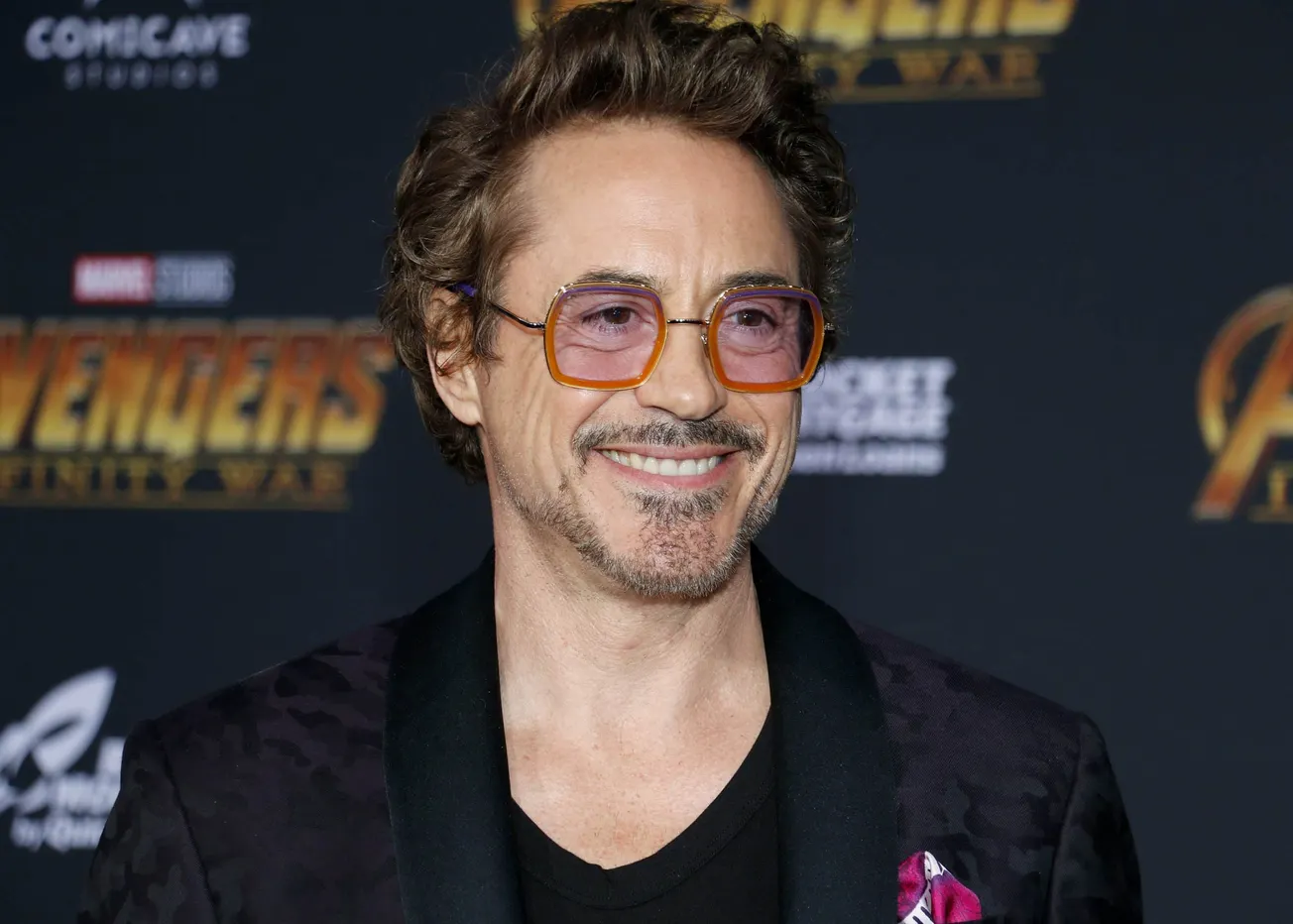 Robert Downey Jr. smiling at an Avengers/ Marvel press event. He has dark facial hair and wears red glasses