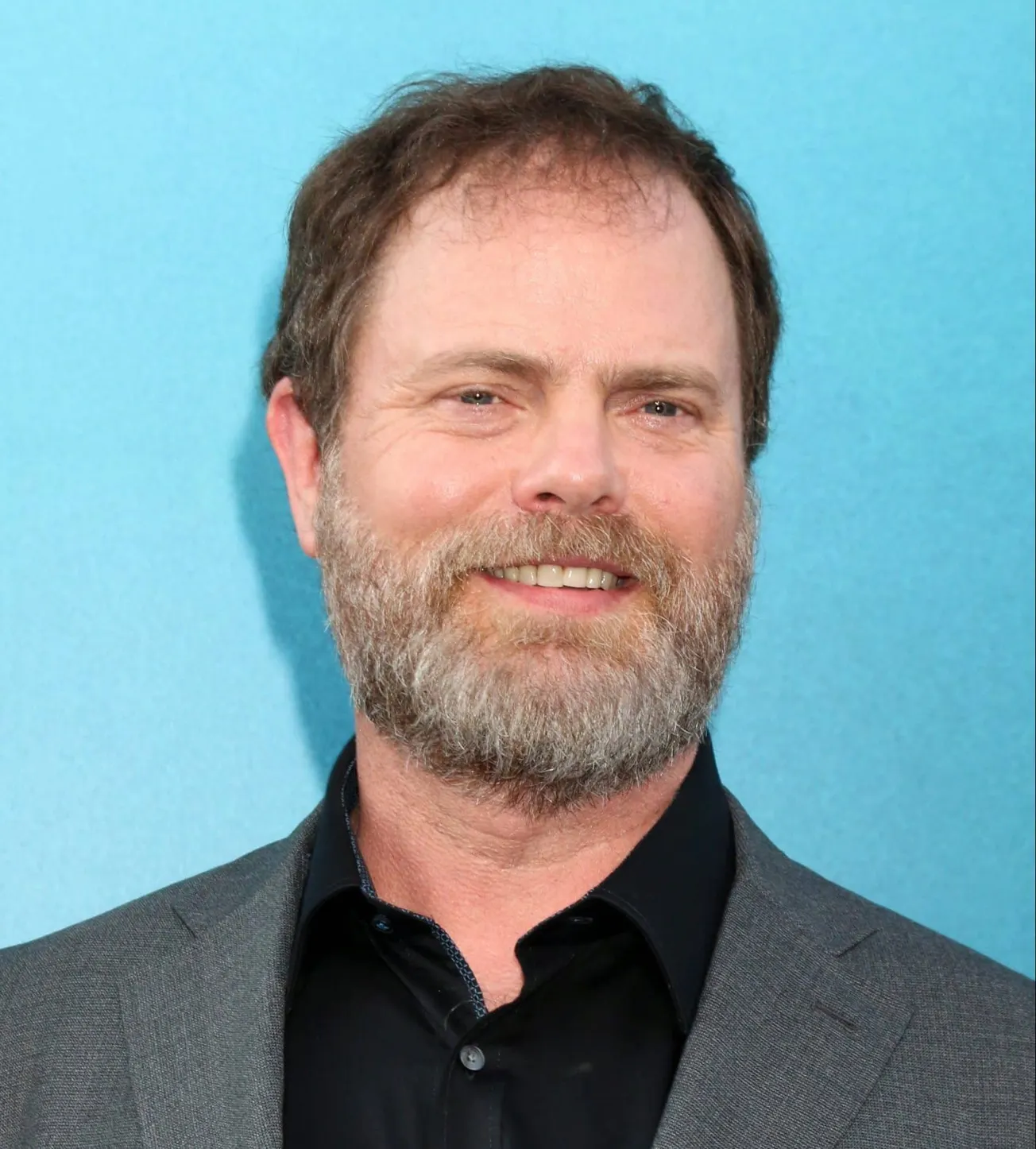 a close up photo of rainn wilson smiling at a press event stnading in front of a blue background