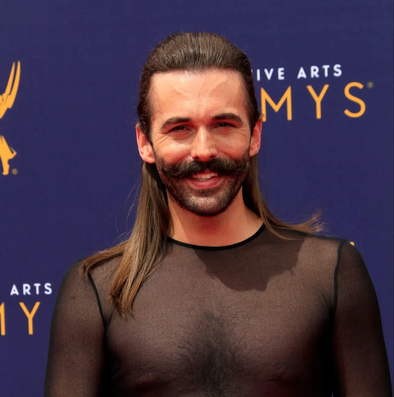 Jonathan Van Ness smiles at a red carpet event. He wears a sheer black top. His hair is half up, slicked back in a pony tail.