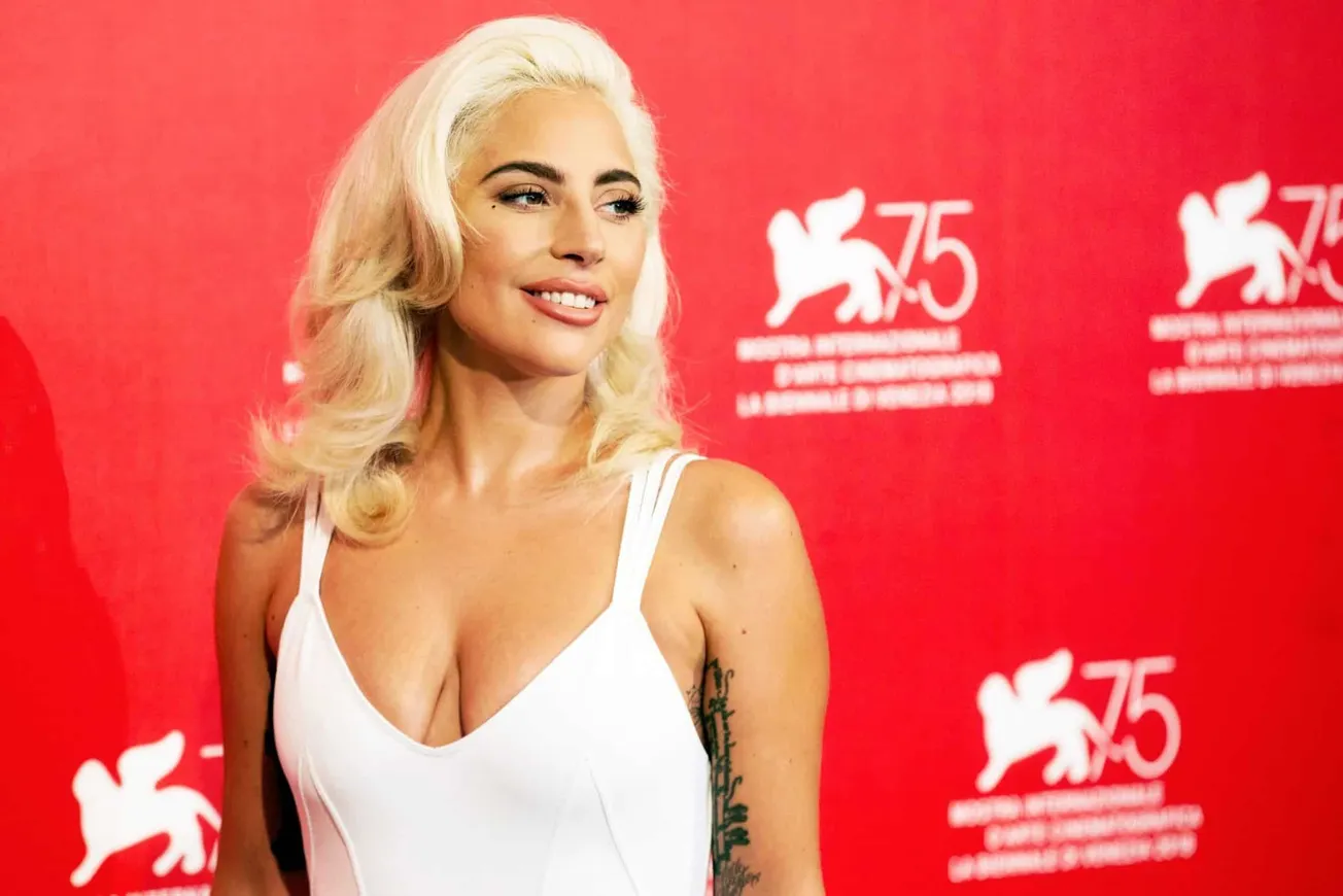 Lady Gaga wears a white dress and has platinum blonde hair that sits elegantly on her shoulders. The background is red. She i