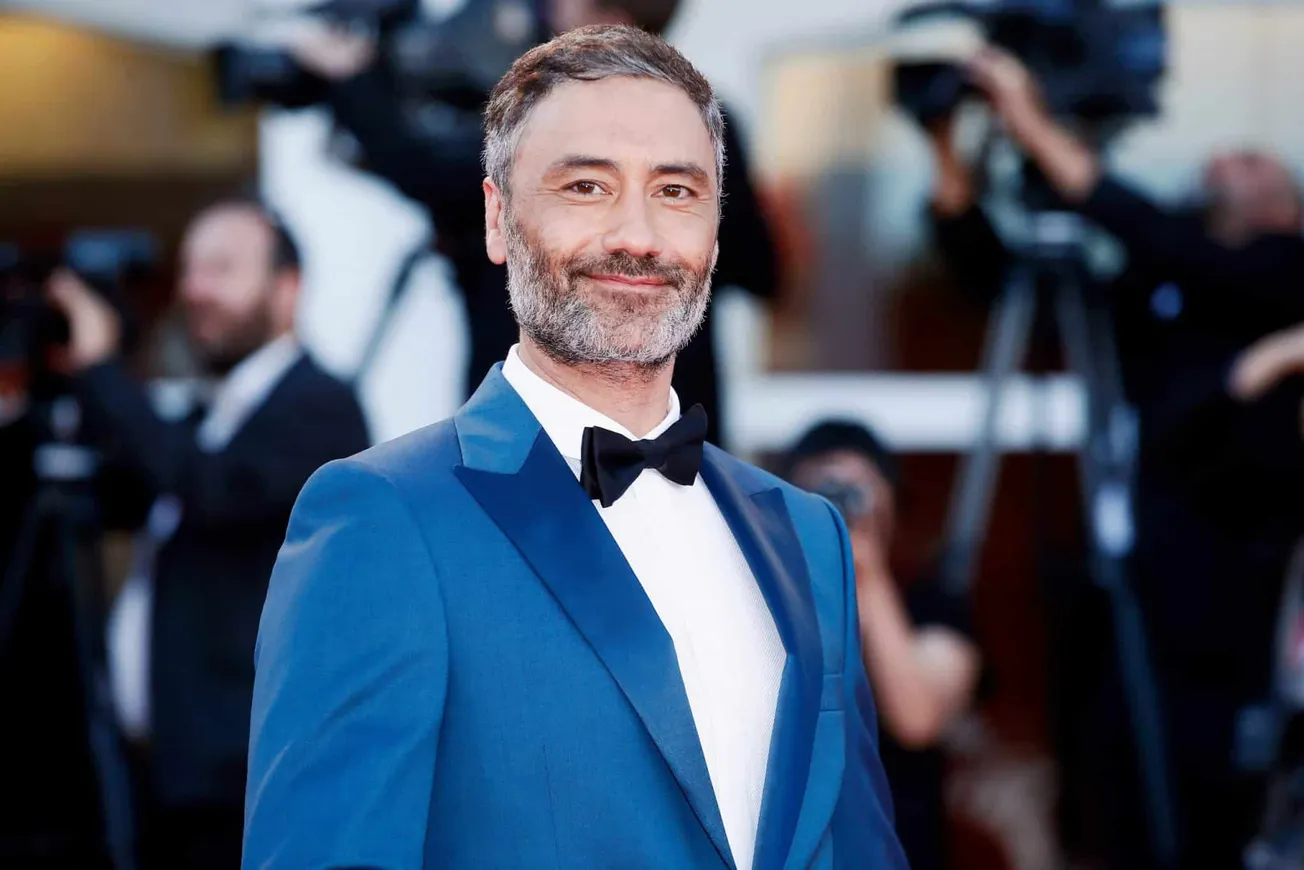 Taika Waititi wears a blue tux and black bow tie at a red carpet event. He has some greying facial hair and is smiling at the