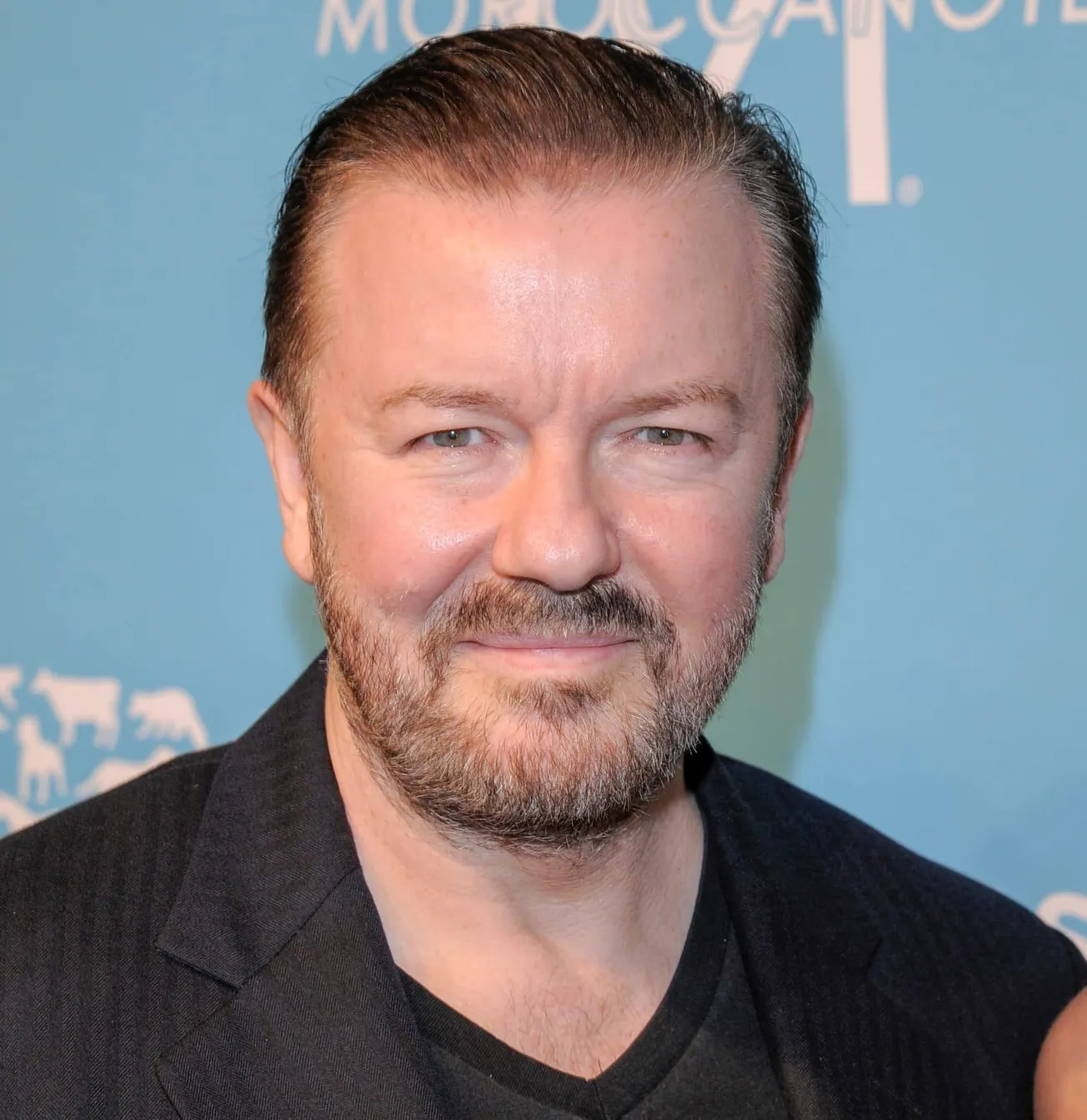 Ricky Gervais smiles at a press event wearing a black suit.