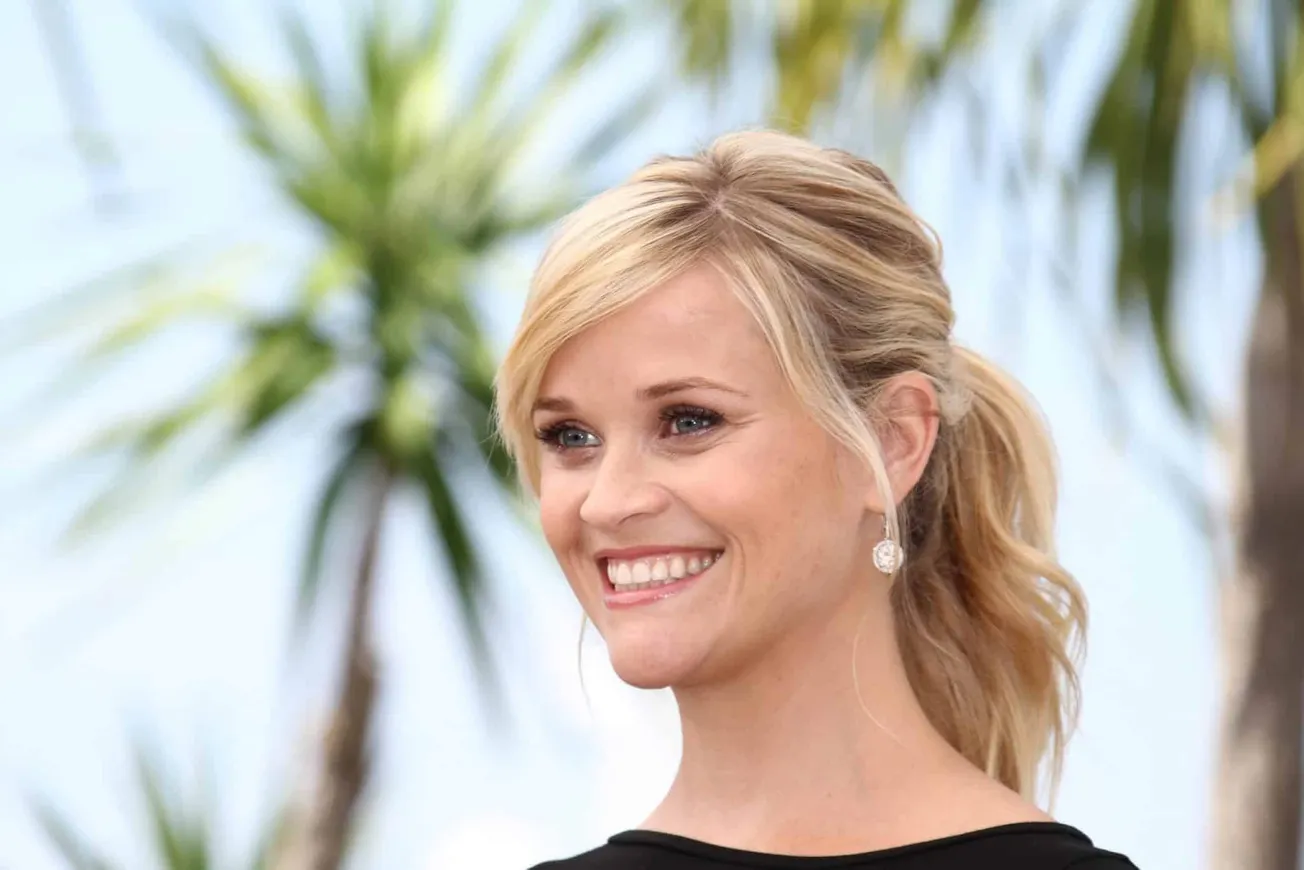 Reese Witherspoon smiles at an outdoor press event. She has blonde hair tie back in a loose ponytail.