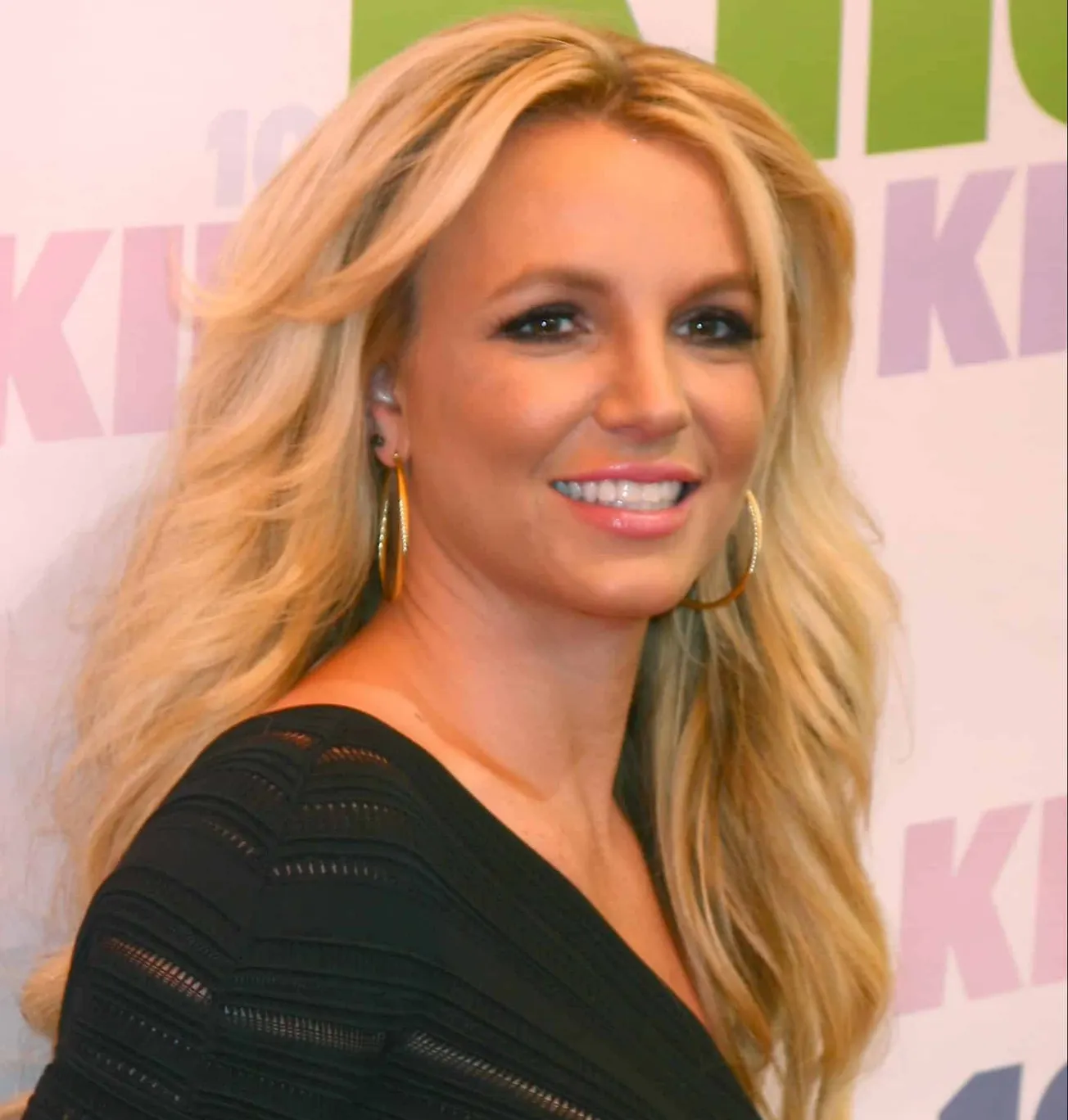 Britney Spears smiles at a red carpet event. She has long blonde layered hair and wears hoop earrings and a black top.
