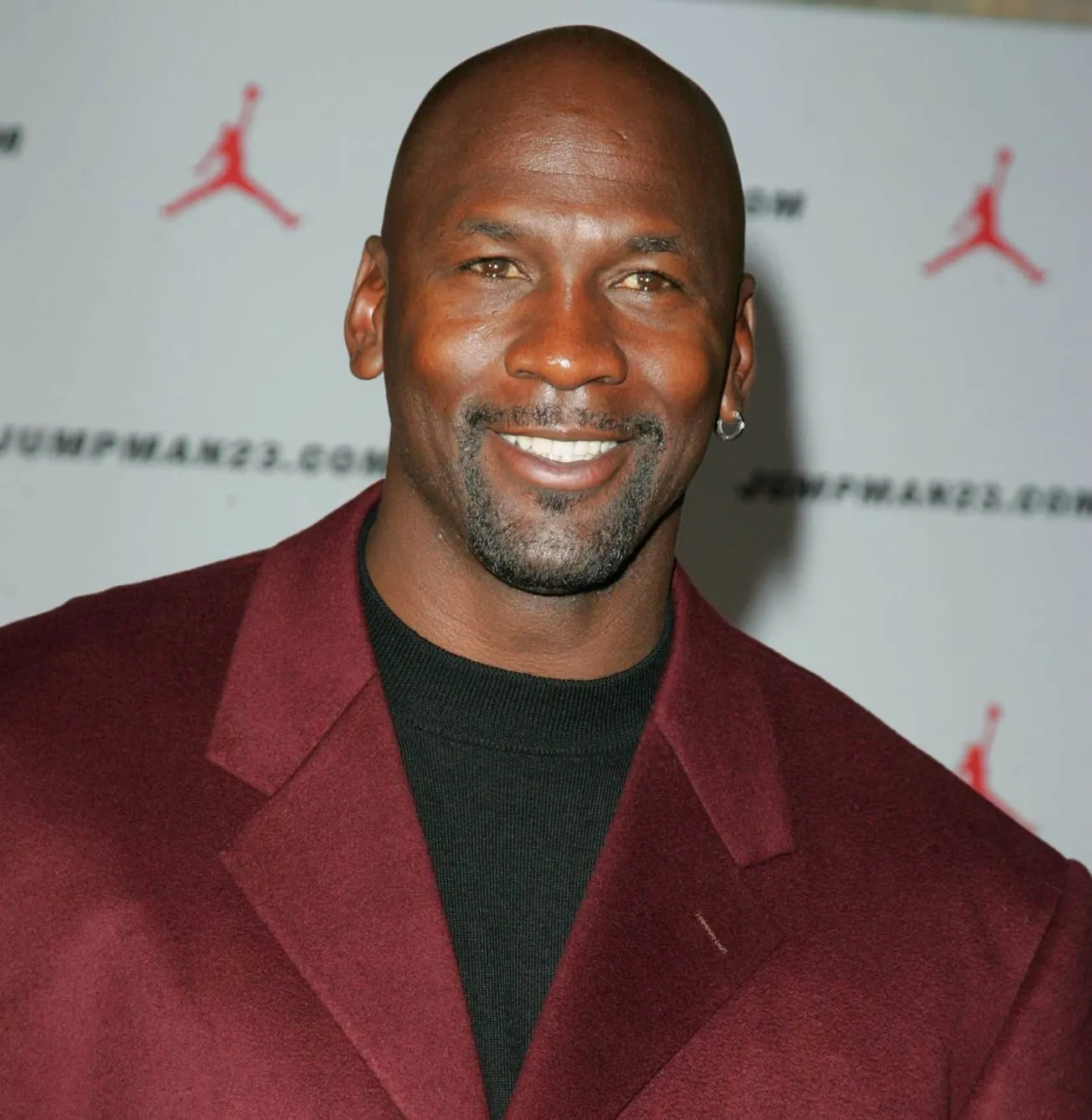 Michael Jordan smiles at a red carpet event. He wears a burgundy suit with a black t-shirt.