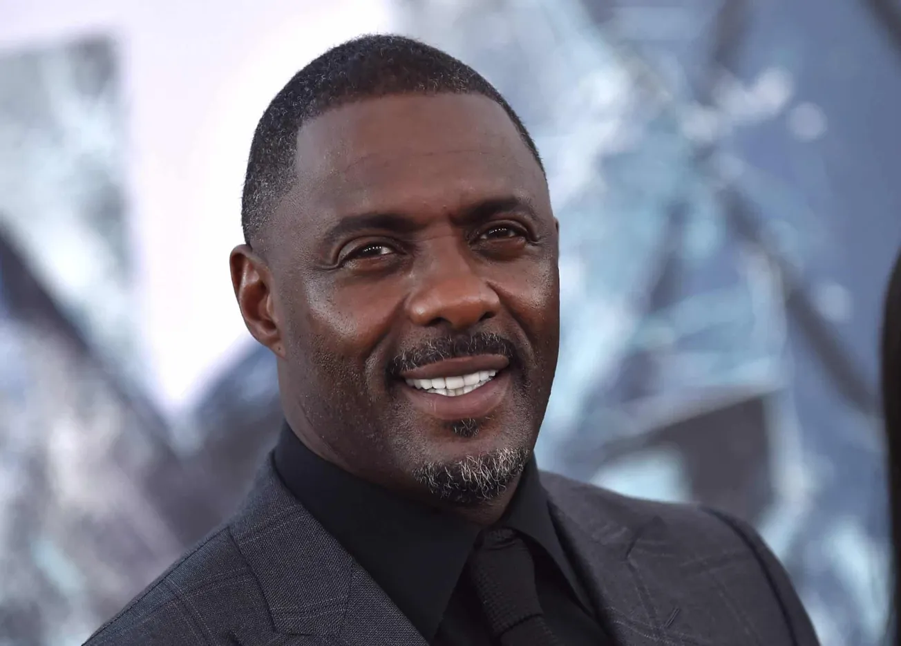 Idris Elba smiles at a red carpet event. He wears a black suit and shirt.