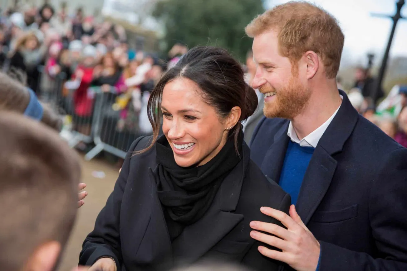 Duke and Duchess of Sussex shake hands with fans at a public event. She wears a dark coat and his her hair tied back. He wear