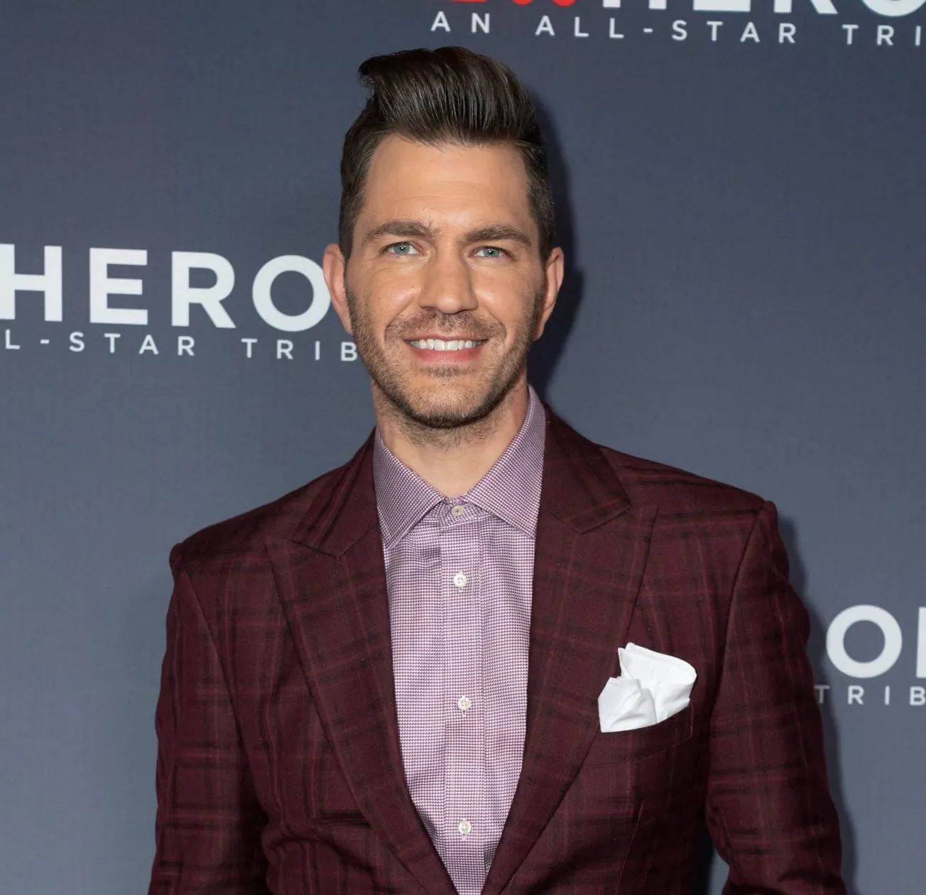 Andy Grammer smiles at a red carpet event. he wears a deep burgundy suit jacket.