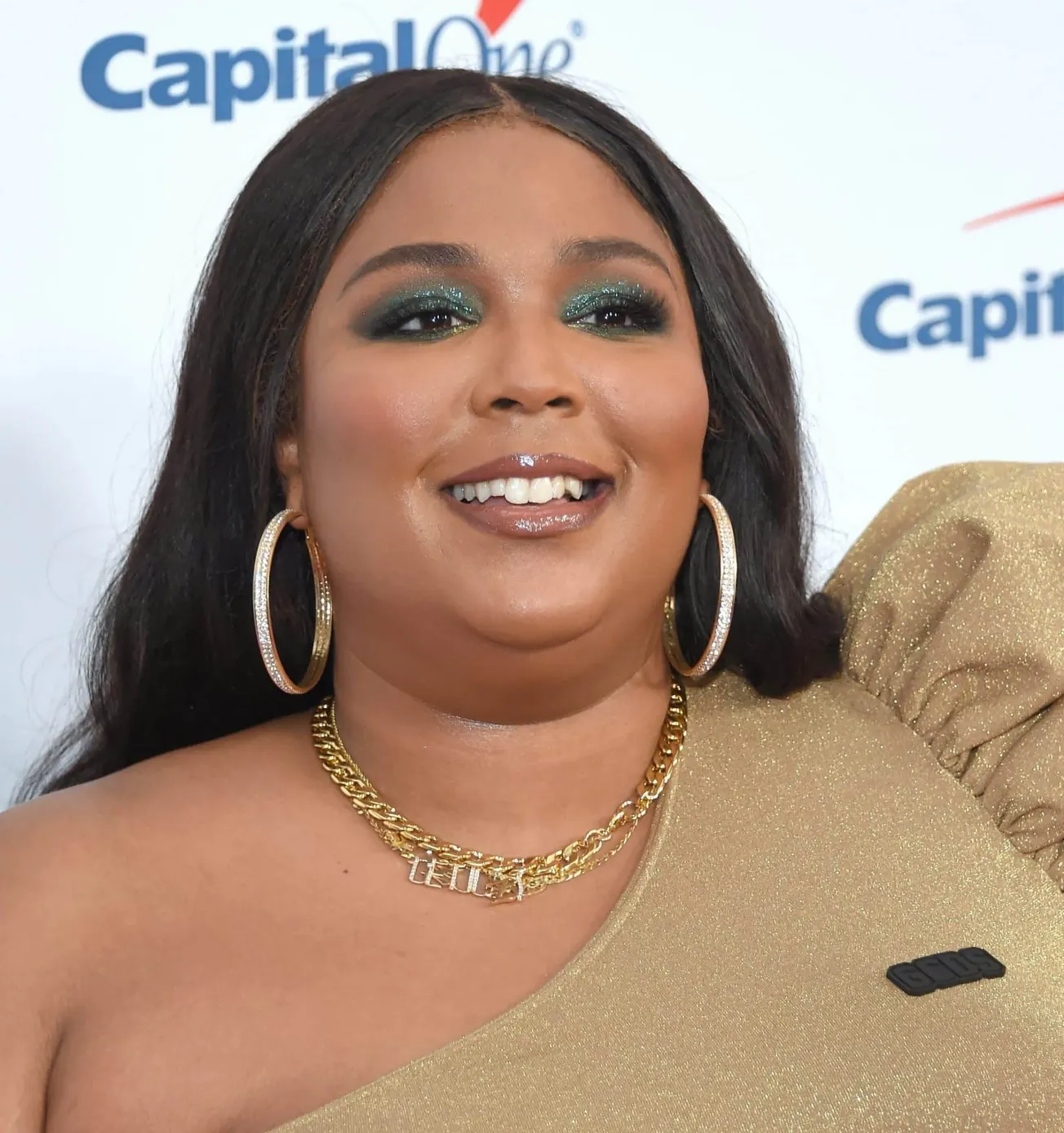 Lizzo smiles at a red carpet event. Her long black hair sits straight, behind her shoulders. She wears big gold hoop earrings