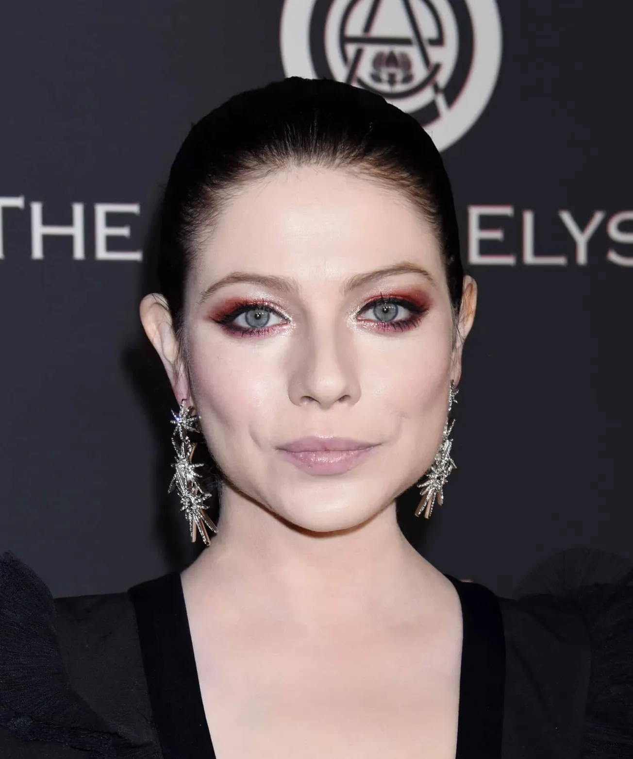 Michelle Trachtenberg smiling at a press event. She has dark hair tied back in a ponytail and wears sparkling earrings.