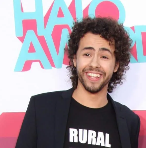 Ramy Youssef smiles at a red carpet event. He has curly balck hair and wears a black t-shirt and suit jacket.