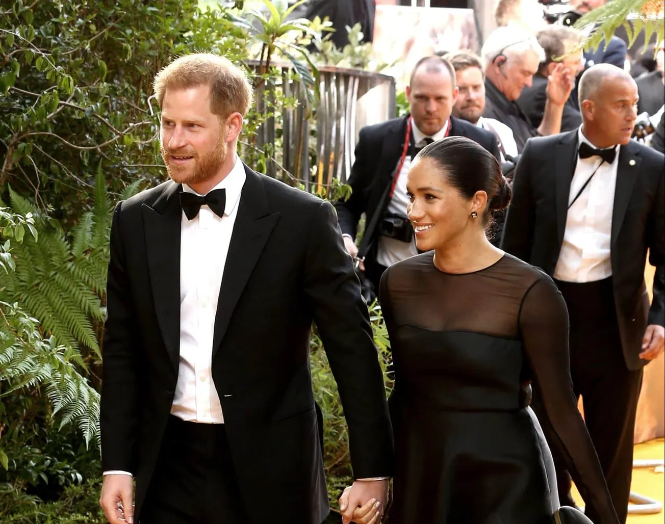 Prince Harry and Duchess Meghan attend a red carpet event, smiling. He wears a tuxedo and she wears a black dress with a shee