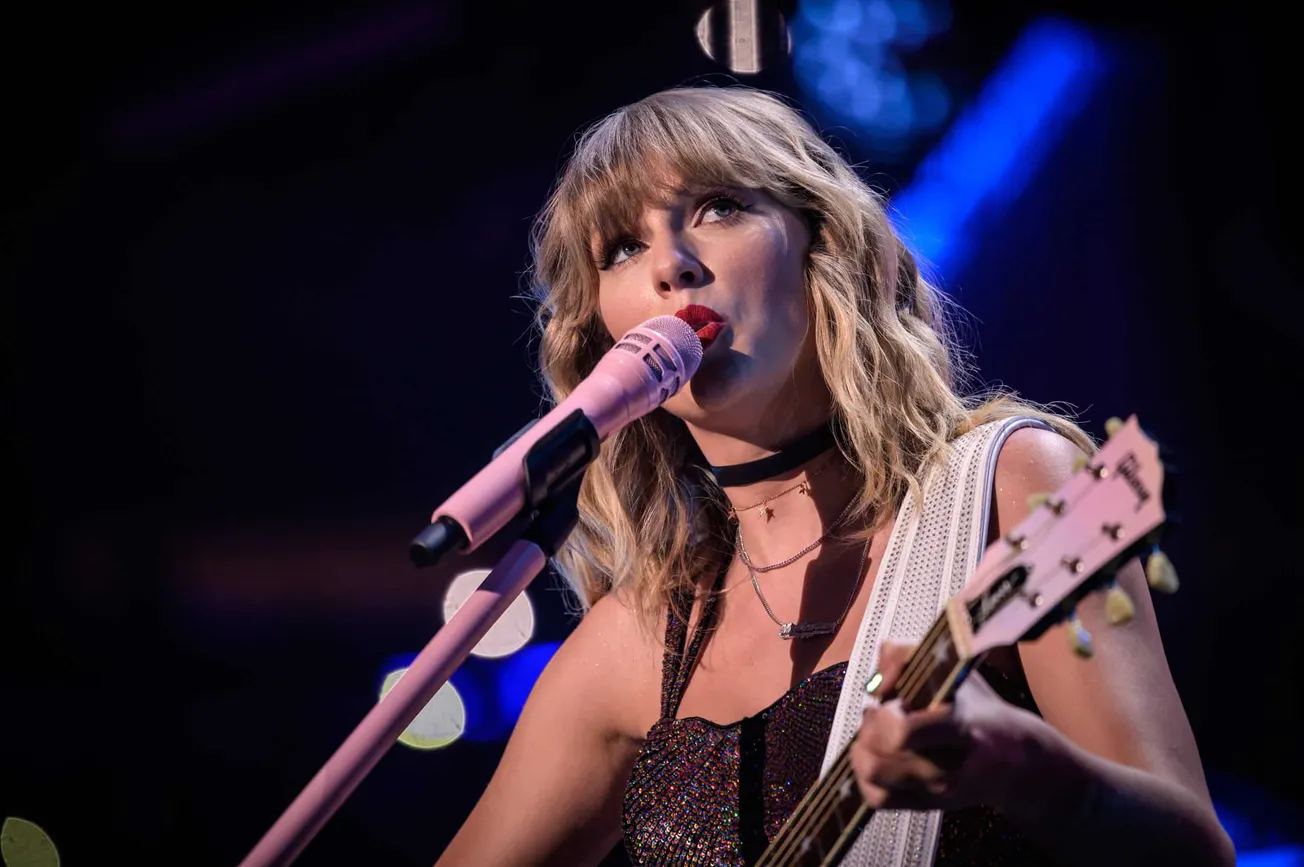 Taylor Swift holding her guitar and standing behind a mic while performing on stage