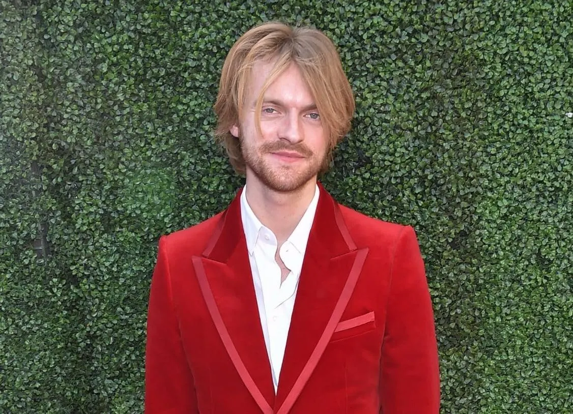 Finneas smiling in a red jacket at a press event