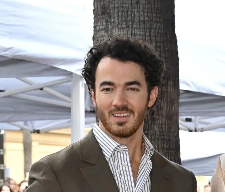 A close up photo of Kevin Jonas wearing a brown suit and white shirt. He's smiling