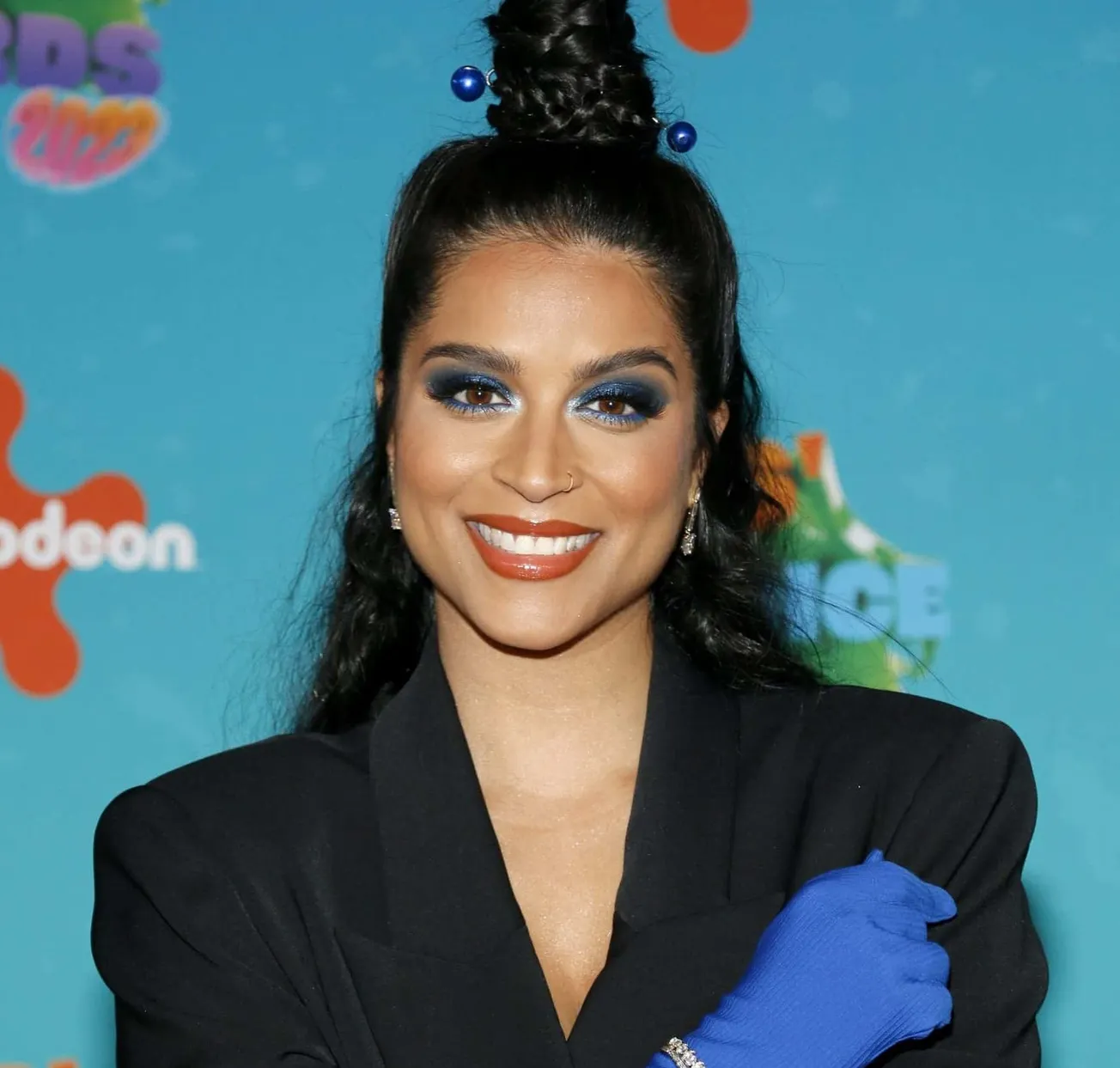 Lilly Singh: Dear Brown Girls video. Lilly smiles at a press event wearing a black suit jacket and blue gloves.