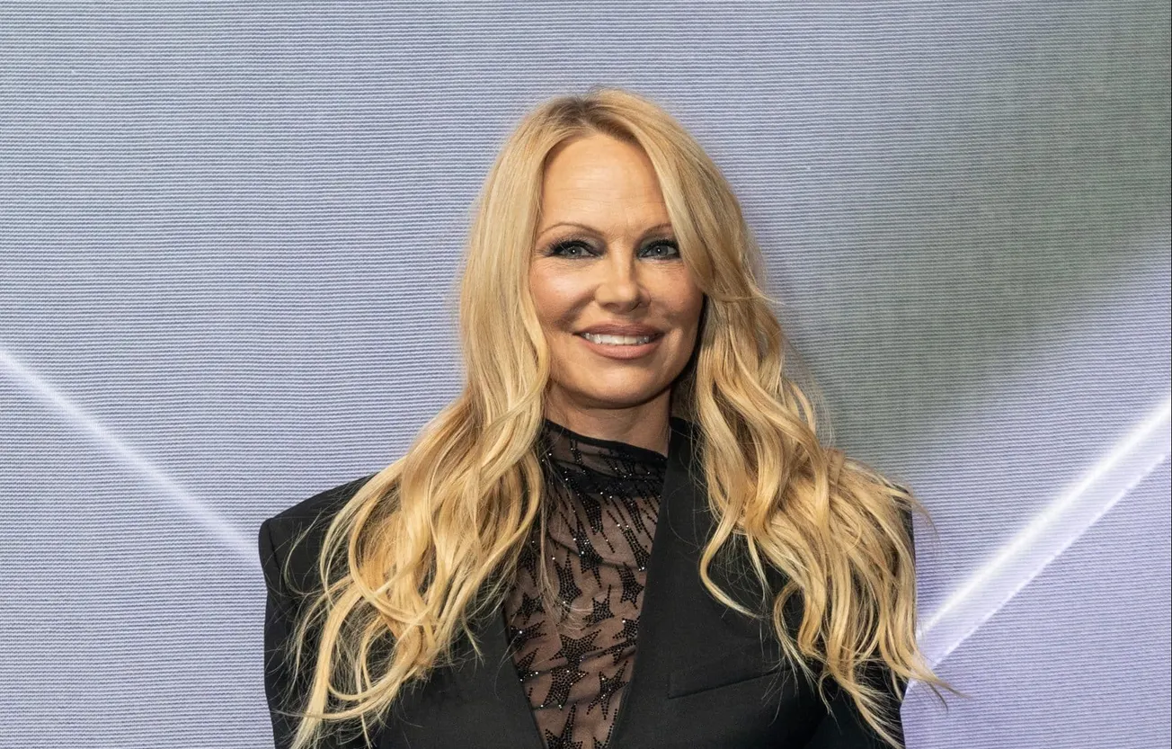Pamela Anderson smiles at a press event wearing a black outfit. Her long blonde hair cascades over her shoulders.