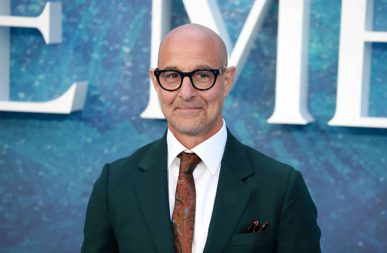 Stnaley Tucci wears a suit and his signature black rimmed glasses. He's smilint at a press event.