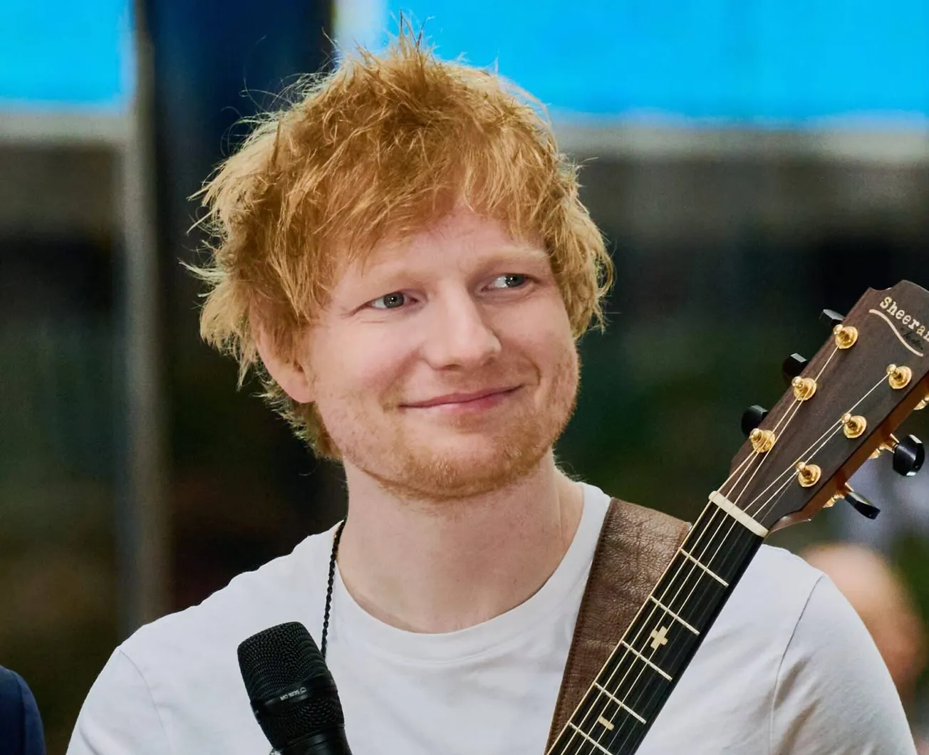 Ed Sheeran holds his guitar and a mic while standing on stage.