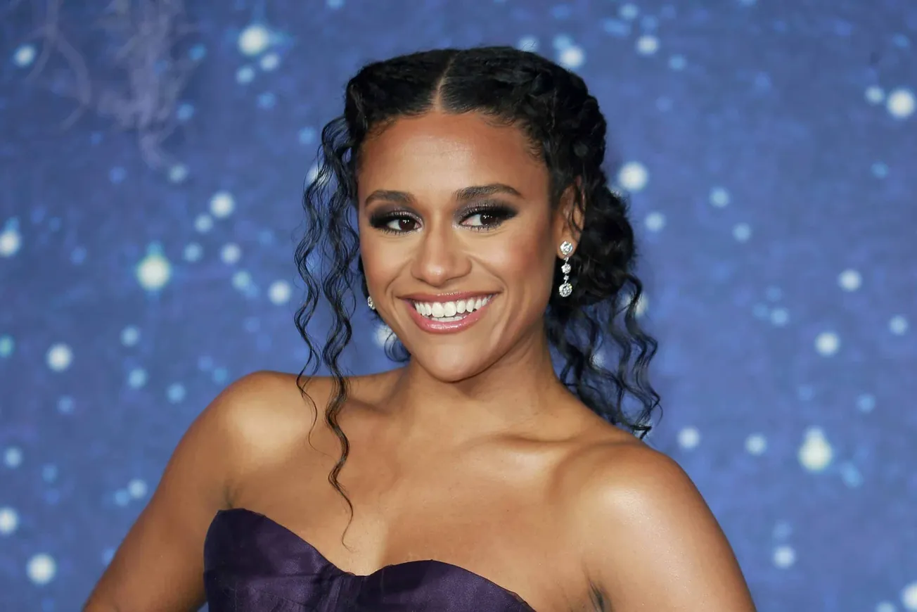 A close up of Ariana DeBose wearing a strapless dress and smiling with a sparkling blue wall behind her. She's at the 'Wish'