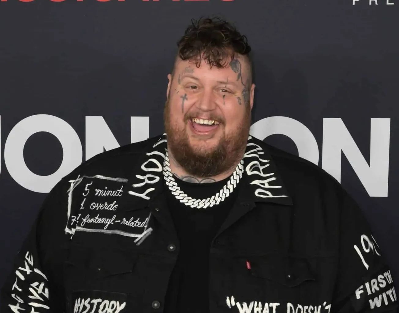 a photo of Jelly Roll smiling. He is dressed in black and has tattoos on his face.