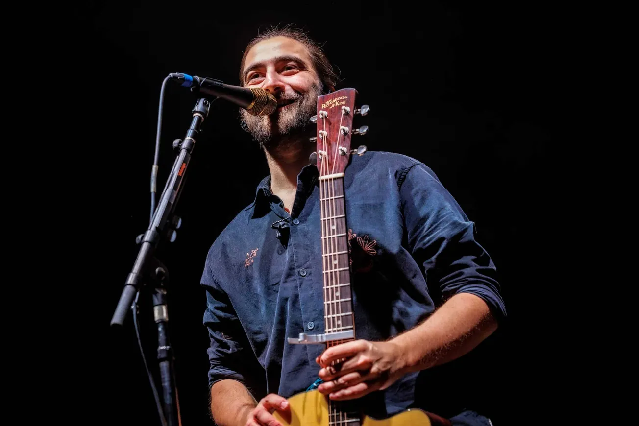 Noah Kahan smiles on stage while holding his guitar