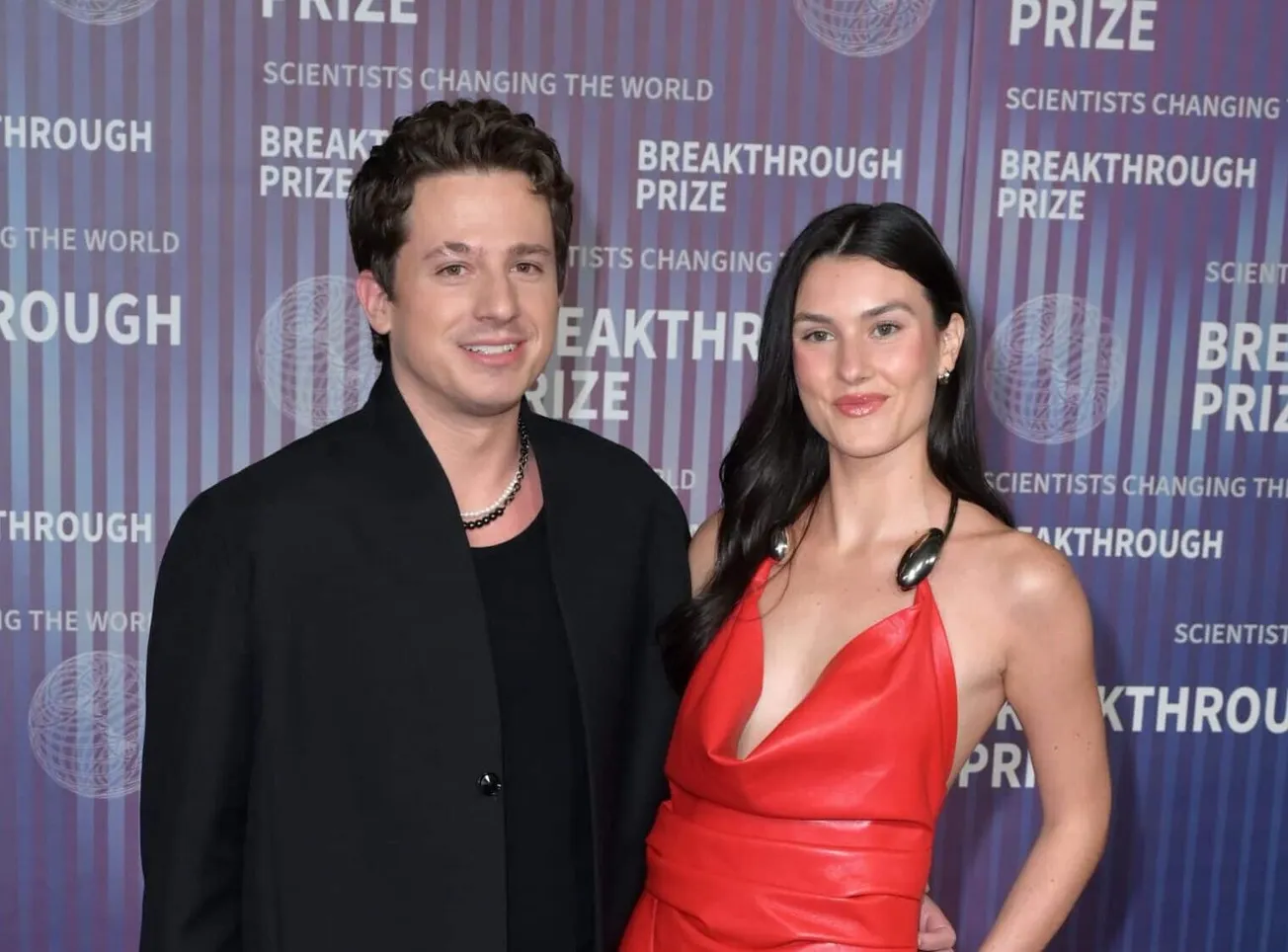 Charlie Puth and Brooke Sansone smile together at a red carpet event. Charlie wears a black suit and Brooke wears a red gown.