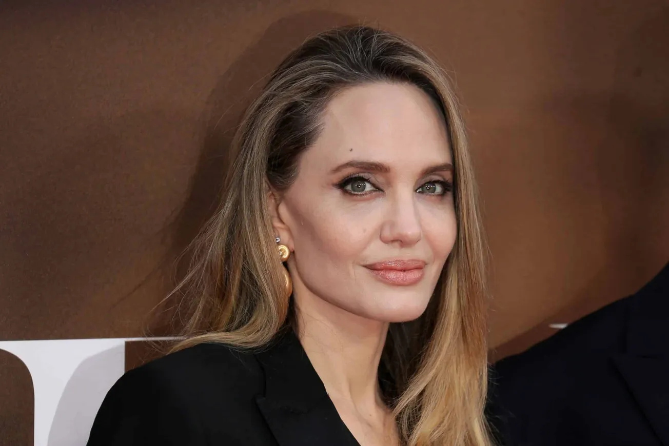 Angelina Jolie smiles at a press event. She has long brown hair and wears a dark suit.