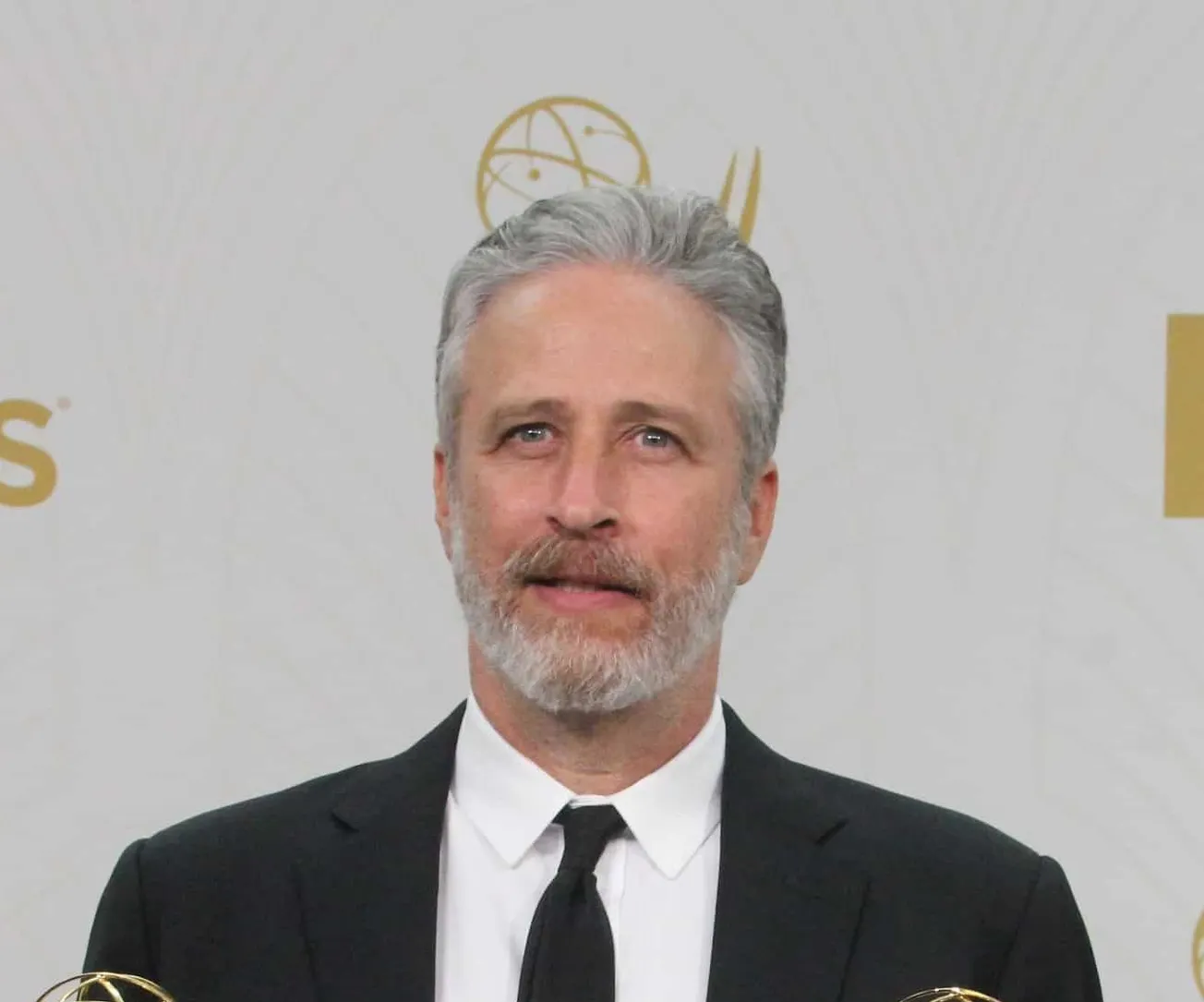 Jon Stewart at an awards event. He has greying hair and a short beard, and wears a suit.