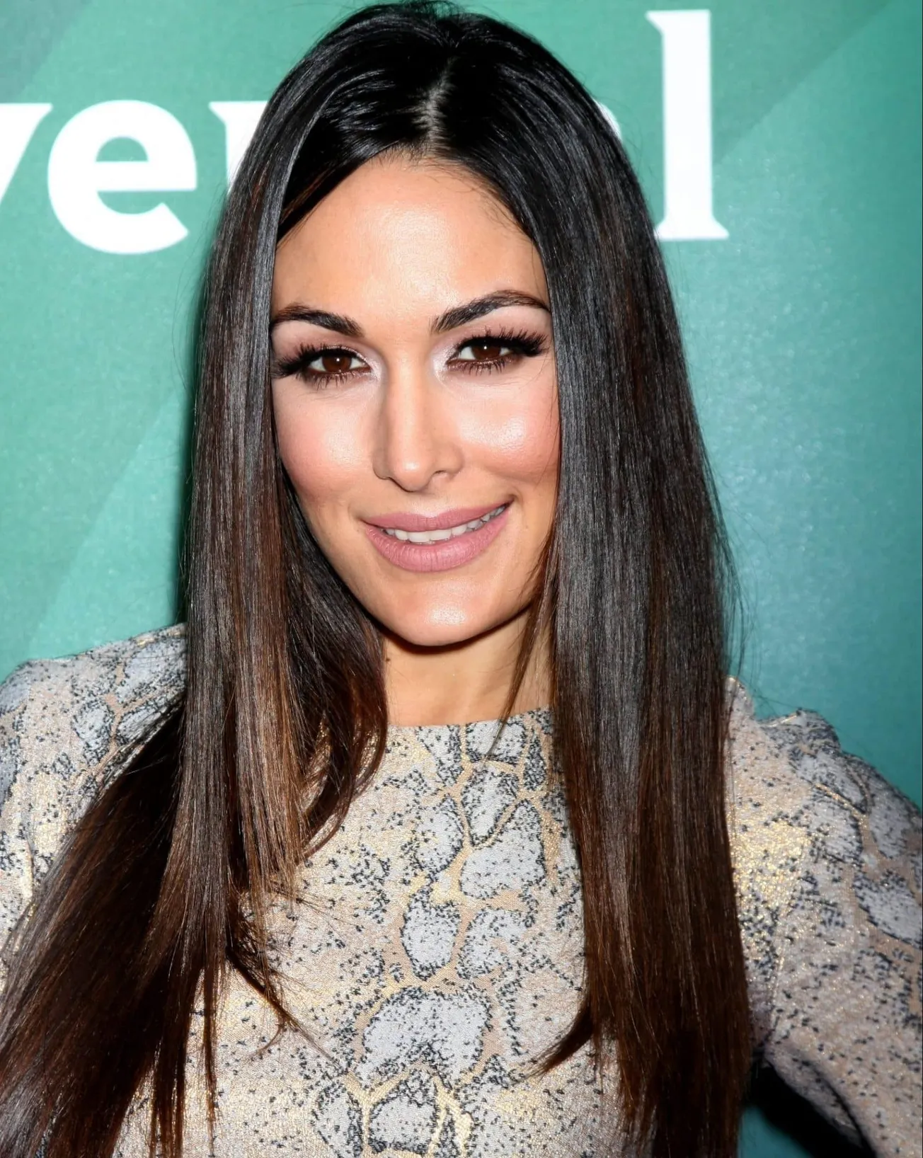 Brie Bella smiles at a red carpet event. She has long straight brown hair and wears a dress that resembles light snakeskin.
