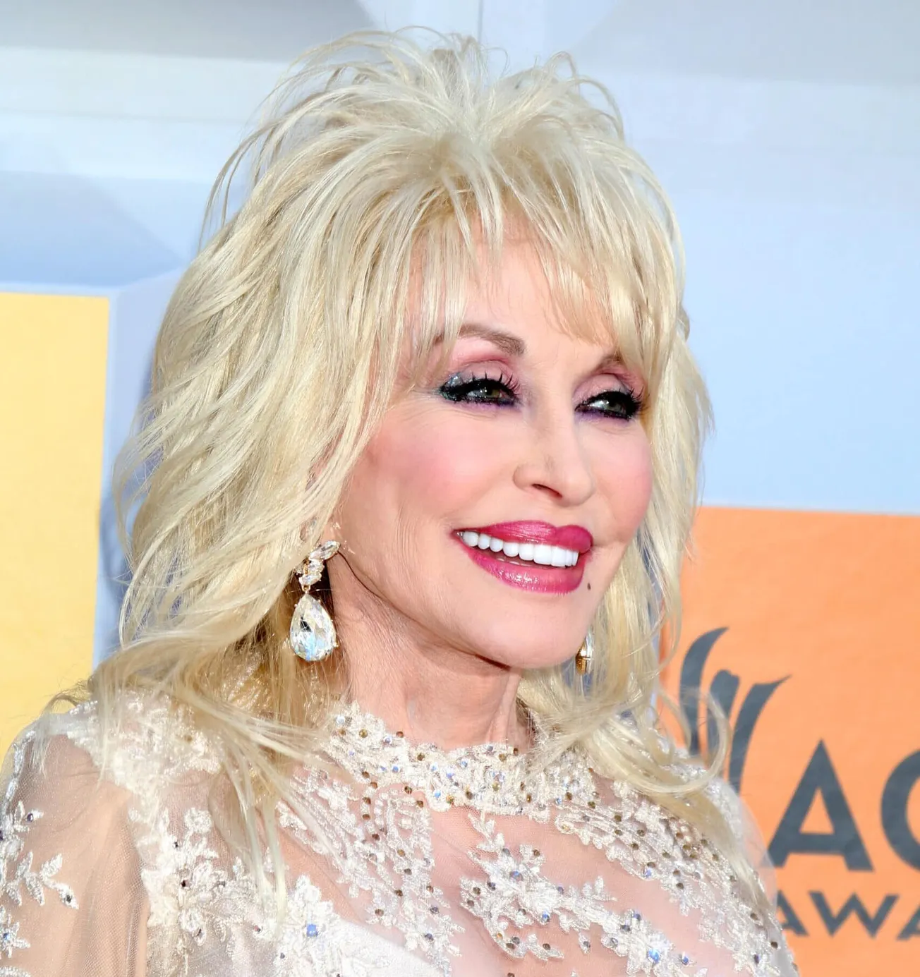 Dolly Parton smiling at a press event. She has big blonde hair and wears bright red lipstick.
