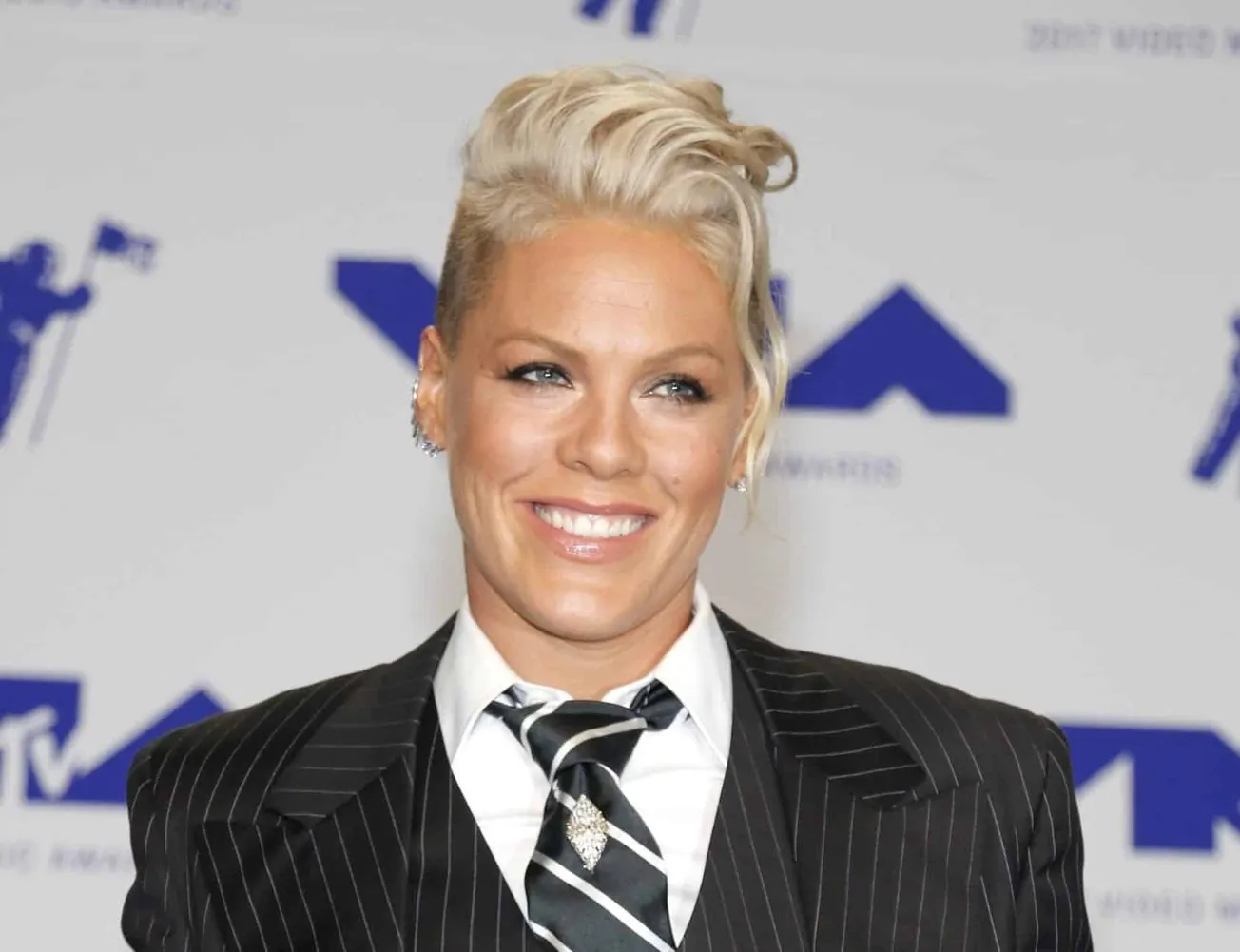 Pink smiles at a red carpet event. She has short blonde hair and wears a black suit and striped tie with a white shirt.