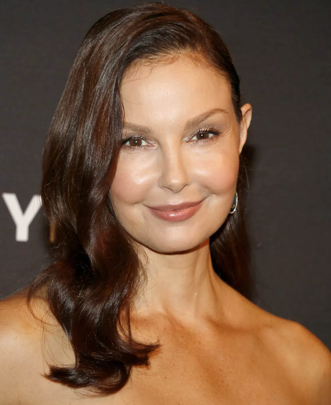 A close up photo of Ashley Judd smiling at a press event.She wears a strapless black dress and has her hair over one shoulder